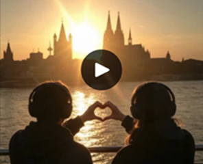 Zwei Menschen sitzen im Sonnenuntergang vor dem Rhein und Kölner Skyline mit Dom und Formen mit ihren Händen ein Herz.