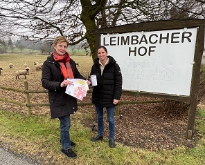 Christel Weyhofen und ihre Tochter Christina Goos mit großen Geldscheinen und einer PalliLev-Spendendose vor dem Hof-Schild des Leimbacher Hofes.