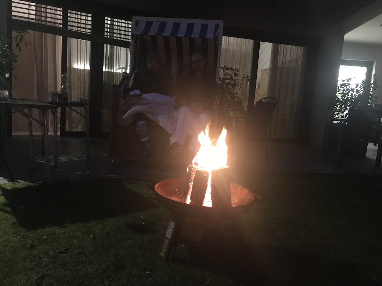 Im Vordergrund brennt ein Lagerfeuer in einer Feuerschale, im Hintergrund sitzen zwei Menschen in Decken gewickelt in einem Strandkorb.