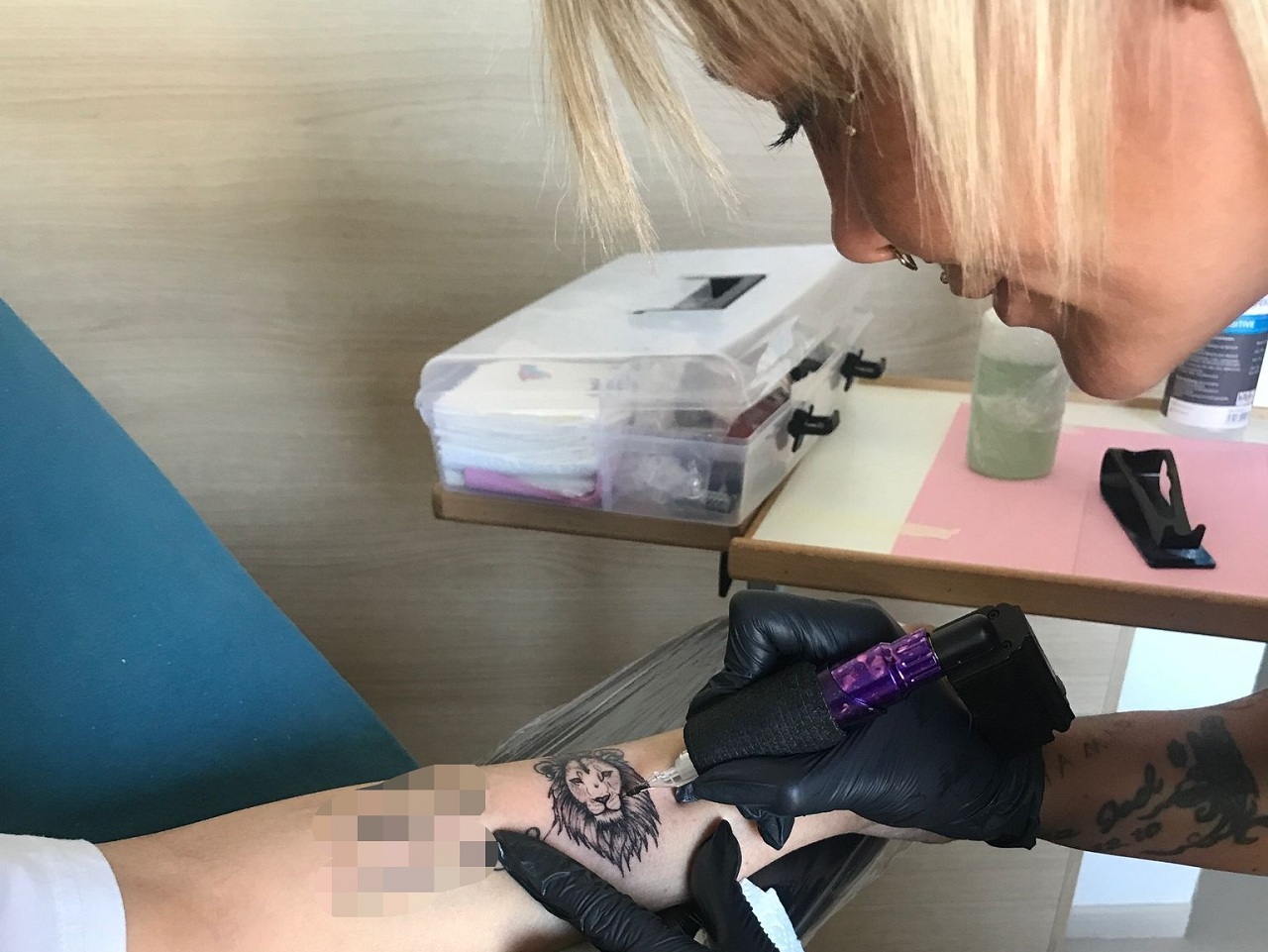 Ein Unterarm, auf dem ein verpixeltes Motiv ist und ein Löwenkopf. Die Tätowiererin beugt sich über den Arm und vervollständigt das Bild. Ihre Ausrüstung steht auf dem Nachttisch.